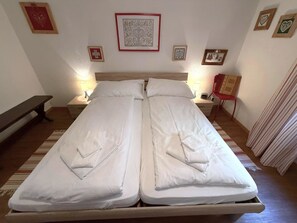A double bed with white linens, two bedside tables with lamps, a bench, and framed pictures on the wall. - Lärchenwald 1808 # 4-bed apartment, Bad, WC 4-Bettwohnung, Bad, WC by Interhome (Leukerbad)