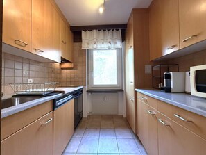 A narrow kitchen with wooden cabinets, a dishwasher, a microwave, and a window with a curtain. - Lärchenwald 1808 # 4-bed apartment, Bad, WC 4-Bettwohnung, Bad, WC by Interhome (Leukerbad)