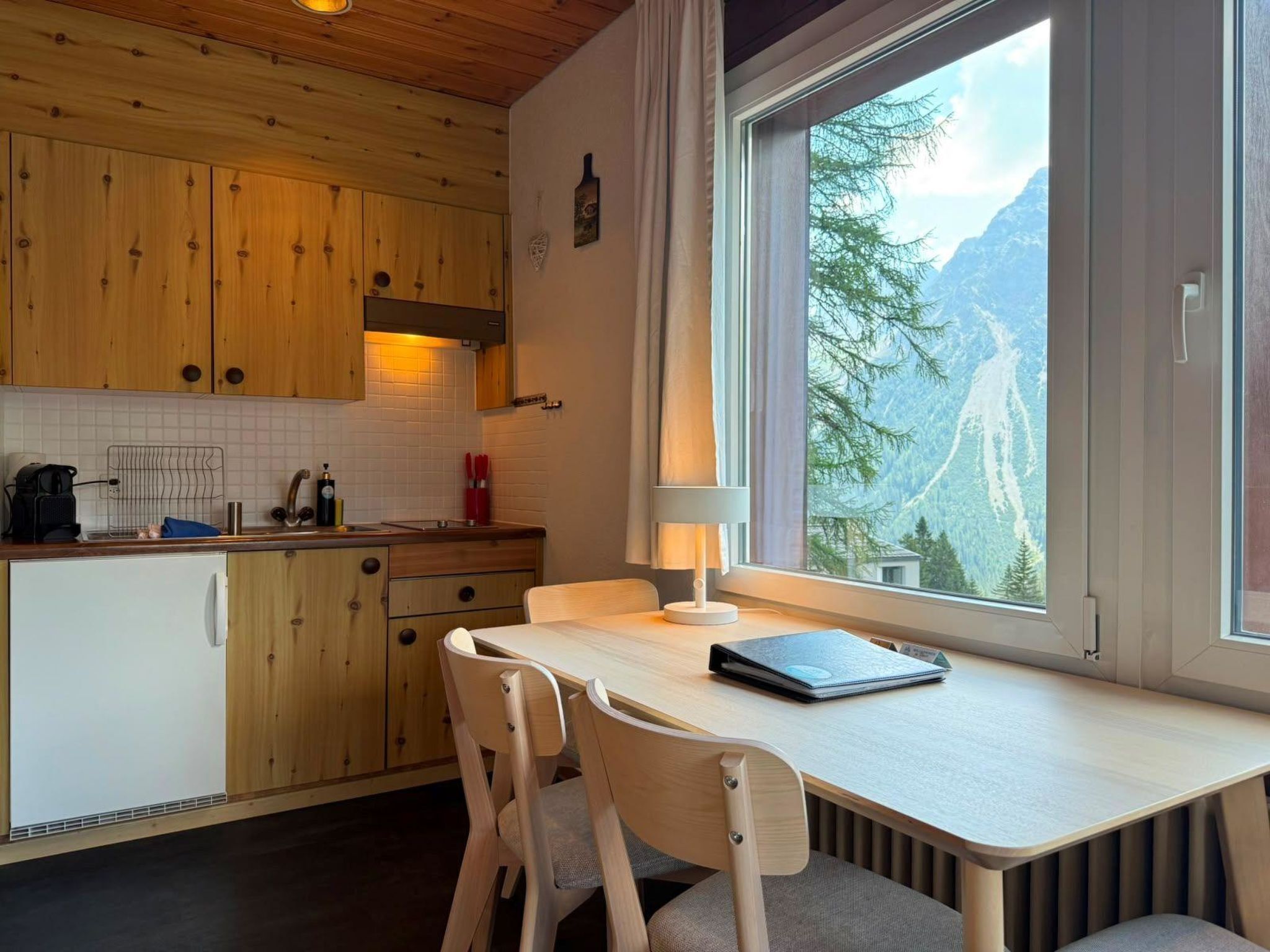 Una cocina con armarios de madera, una mesa de comedor con sillas, una ventana con vista a la montaña y una lámpara sobre la mesa.