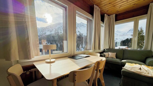 Una stanza accogliente con soffitto in legno, un tavolo da pranzo con sedie, un divano e ampie finestre con vista sulle montagne.