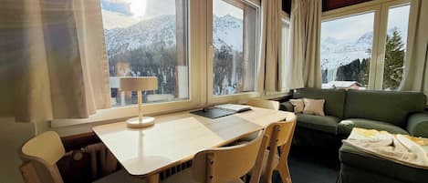A cozy room with a wooden ceiling, a dining table with chairs, a sofa, and large windows offering a mountain view.