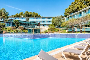 En swimmingpool med solstolar, en byggnad med flera våningar och träd i bakgrunden.