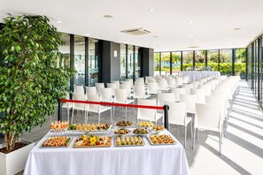 Ett konferensrum med ett vitt bord som är dukat med olika sorters mat, omgivet av vita stolar, och en stor krukväxt.