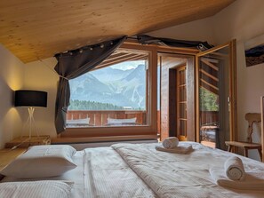 A bedroom with a bed, a nightstand, a lamp, and a view of mountains through a large window. - Furkasunna by Arosa Holiday Appartement/Fewo, Dusche oder Bad, WC, 7-Bettwohnung by Interhome (Arosa)