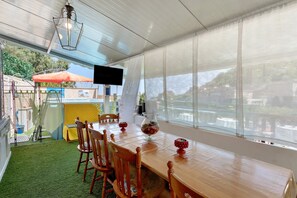 Ein Essbereich mit einem langen Holztisch, Stühlen, einem Fernseher und Blick auf ein Gewässer durch große Fenster.