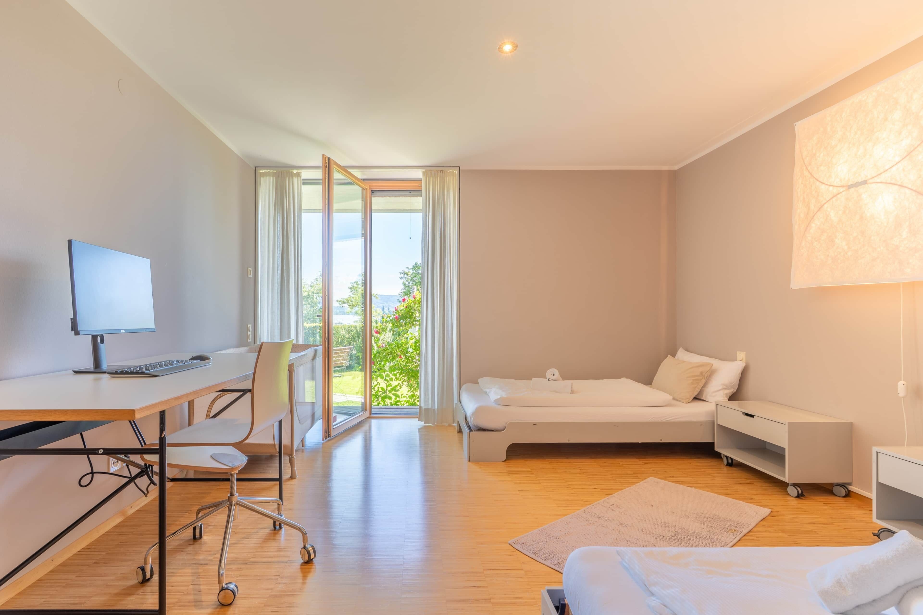 Une chambre d’hôtel moderne équipée d’un bureau, d’un ordinateur, d’une chaise, d’un lit, d’une table de chevet et offrant une vue sur la verdure.