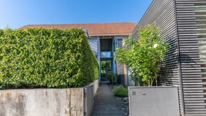 Un étroit sentier mène à un bâtiment moderne doté d’un toit en pente et d’un bac en béton orné de plantes vertes.