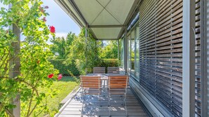 Un espace extérieur abrité, doté d’une terrasse en bois, de chaises en métal et offrant une vue sur un jardin avec des arbustes et un arbre.
