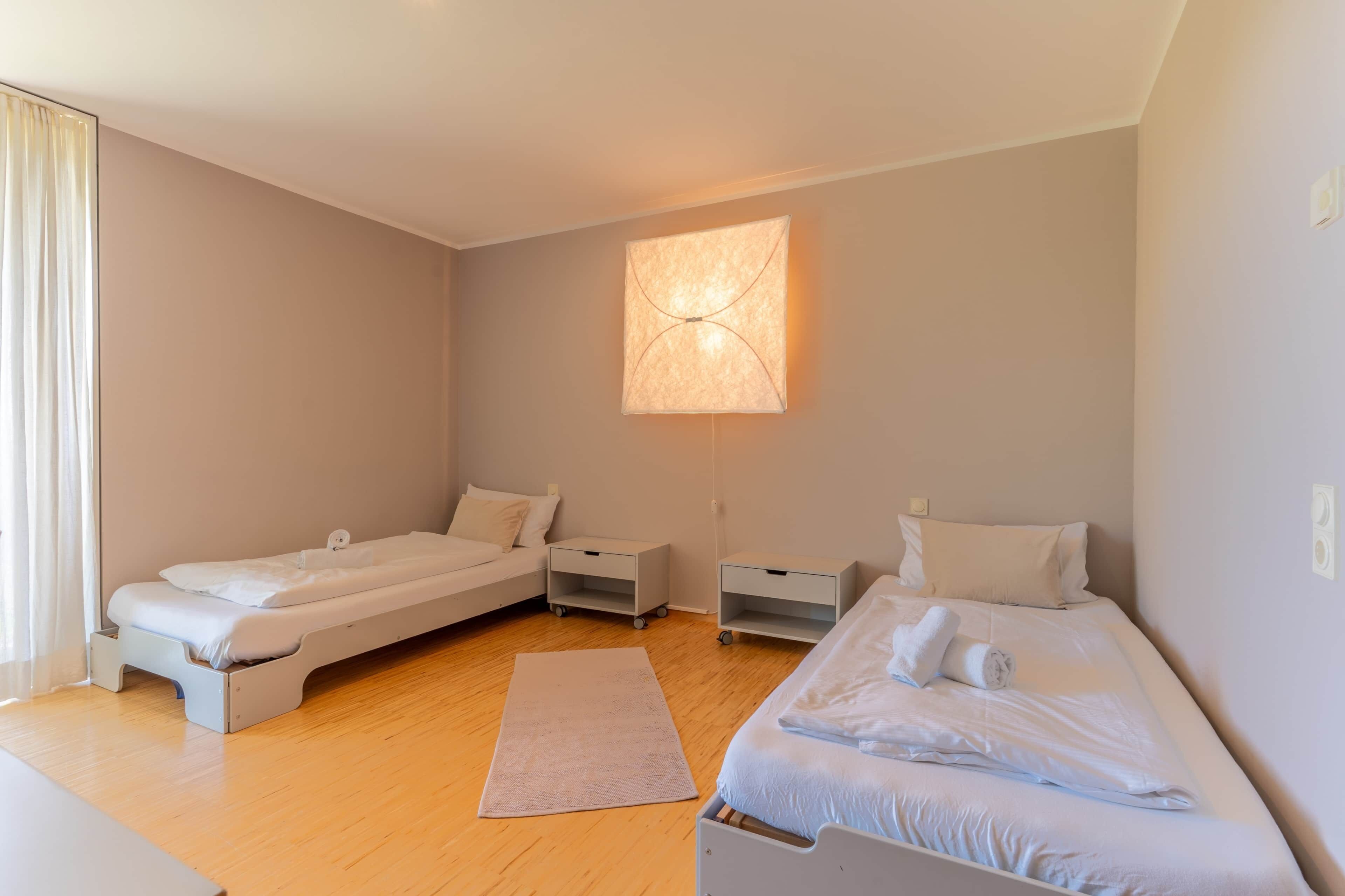 Une chambre avec deux lits simples, une table de chevet, une commode, une lampe fixée au mur et une fenêtre avec des rideaux.