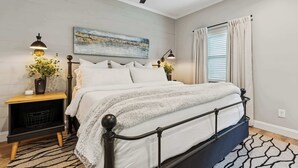 A bedroom with a bed, a ceiling fan, a nightstand with a lamp, a painting on the wall, and a window with curtains. - Loft Haven Rooftop Terrace (Charlotte)