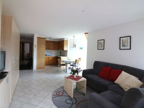 Ein Wohnzimmer mit grauem Sofa, einem kleinen Couchtisch und einem Fernseher links. Im Hintergrund befindet sich eine Küche mit Holzschränken sowie ein Essbereich mit Stühlen.