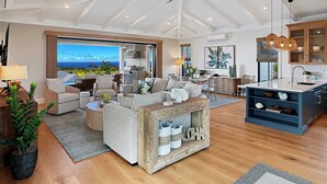 A spacious living room with a large window, a ceiling fan, and a kitchen area with wooden cabinets. - Hali'a Mai Distant Ocean View Koloa Home w/ Pool (Koloa)