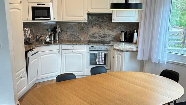 Moderni keittiö, jossa on valkoiset kaapit, puinen ruokapöytä ja mustat tuolit.