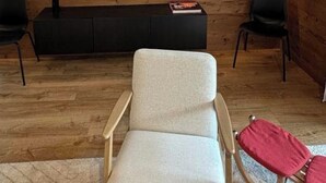 Huoneessa on puinen seinä, tummanvärinen viihdekeskus, jonka yläpuolella on seinälle asennettu taulutelevisio, kaksi mustaa tuolia ja vaaleanvärinen sohva.