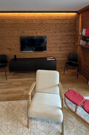 A room with a wooden wall, a flat-screen TV mounted above a dark-colored entertainment unit, two black chairs, and a light-colored sofa. - Edelweiss D 13 # 6-bed apartment, Dusche, WC 6-bed apartment, Dusche, WC by Interhome (Leukerbad)