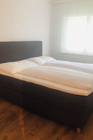 Ein Bett mit dunklem Kopfteil in einem Zimmer mit Holzdielen und einem Fenster mit transparenten Vorhängen.