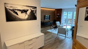 Moderni olohuone, jossa on suuri mustavalkoinen vuoristomaisemamaalaus, valkoinen mediakeskus ja ruokailutila, jossa on lasipöytä ja -tuolit.