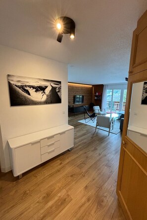 A modern living room with a large black and white mountain landscape painting, a white entertainment unit, and a dining area with a glass table and chairs. - Edelweiss D 13 # 6-bed apartment, Dusche, WC 6-bed apartment, Dusche, WC by Interhome (Leukerbad)