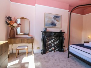 A bedroom with a bed, a bedside table, a dresser, a mirror, a vase with flowers, a fireplace, and a painting on the wall. - Fox House (Llandudno)