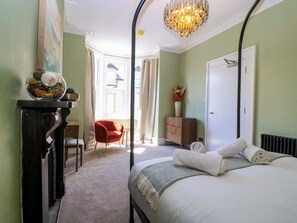 Ein Schlafzimmer mit einem Bett, einem Sessel, einem Schminktisch, einem Schreibtisch und einem Fenster mit Vorhängen.