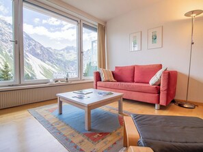 Ein Wohnzimmer mit rotem Sofa, einem Couchtisch, einer Stehlampe und Blick auf Berge durch große Fenster.