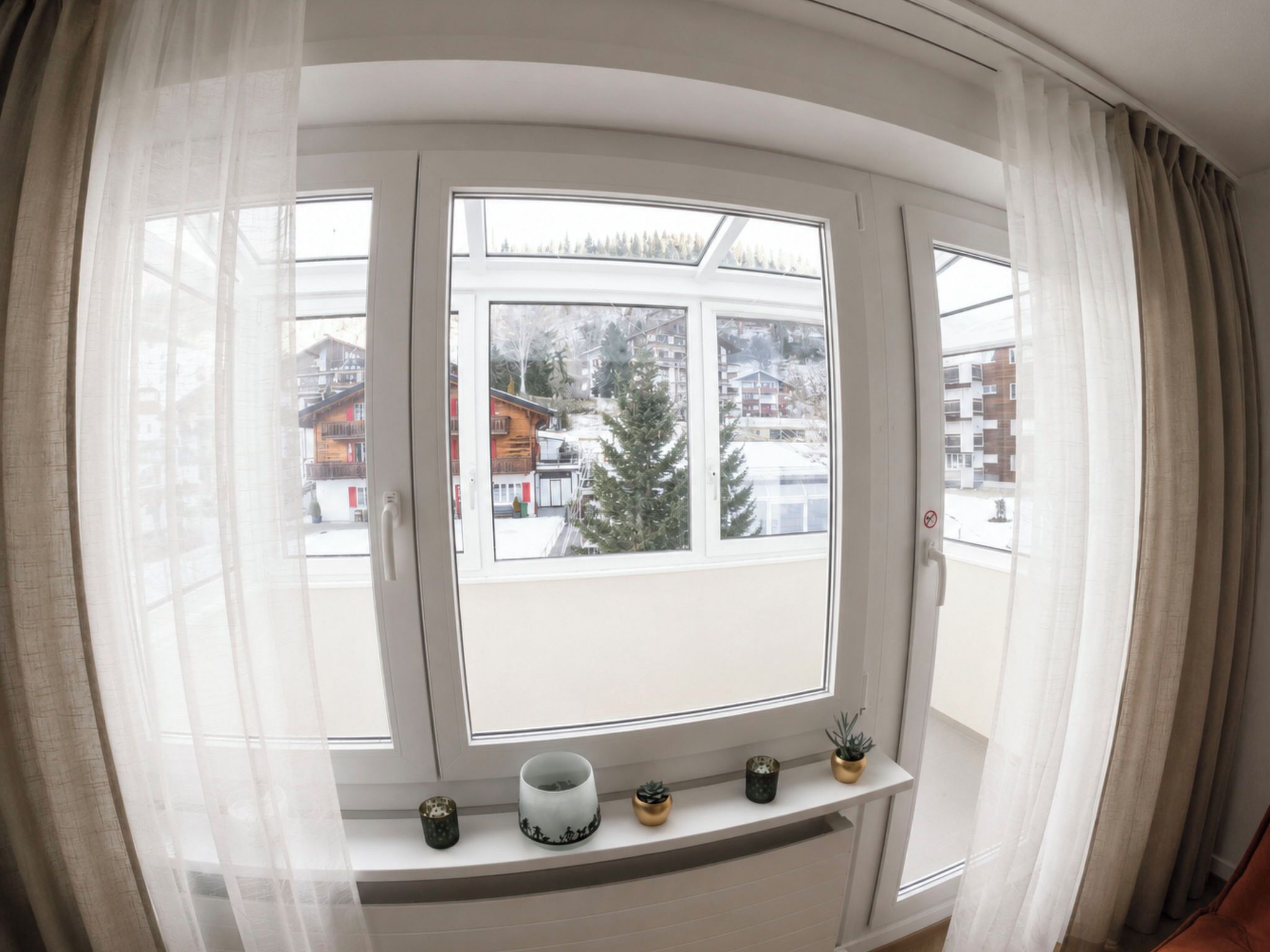 Um quarto com uma janela grande, uma estante branca com objetos decorativos e vista para edifícios e árvores.