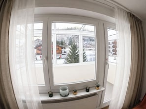 A room with a large window, a white shelf with decorative items, and a view of buildings and trees outside. - Majesta Leukerbad # 2-bed apartment, Dusche, WC by Interhome (Leukerbad)