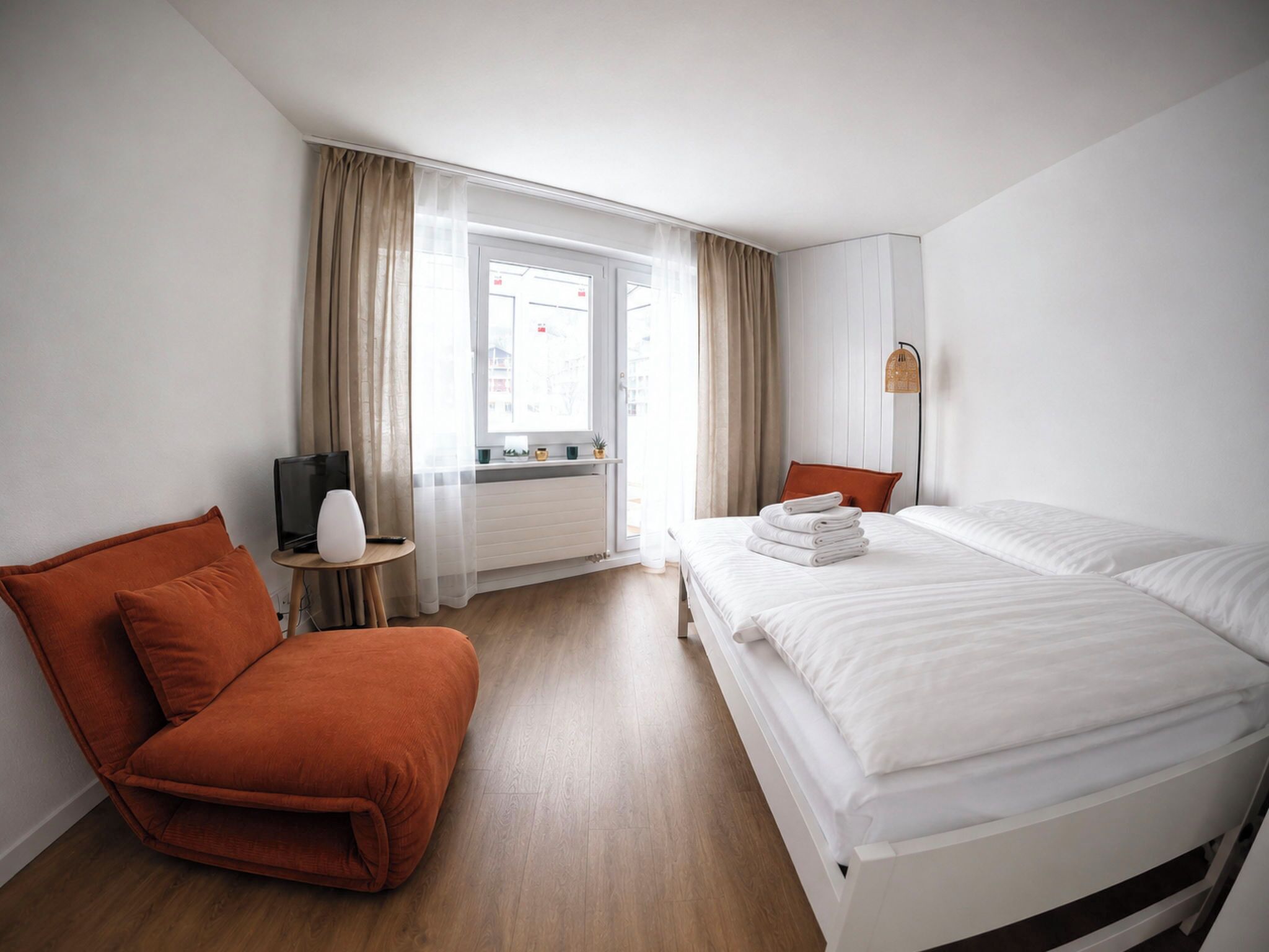 A hotel room with a bed, a chair, a TV, a small table, and a window with curtains.
