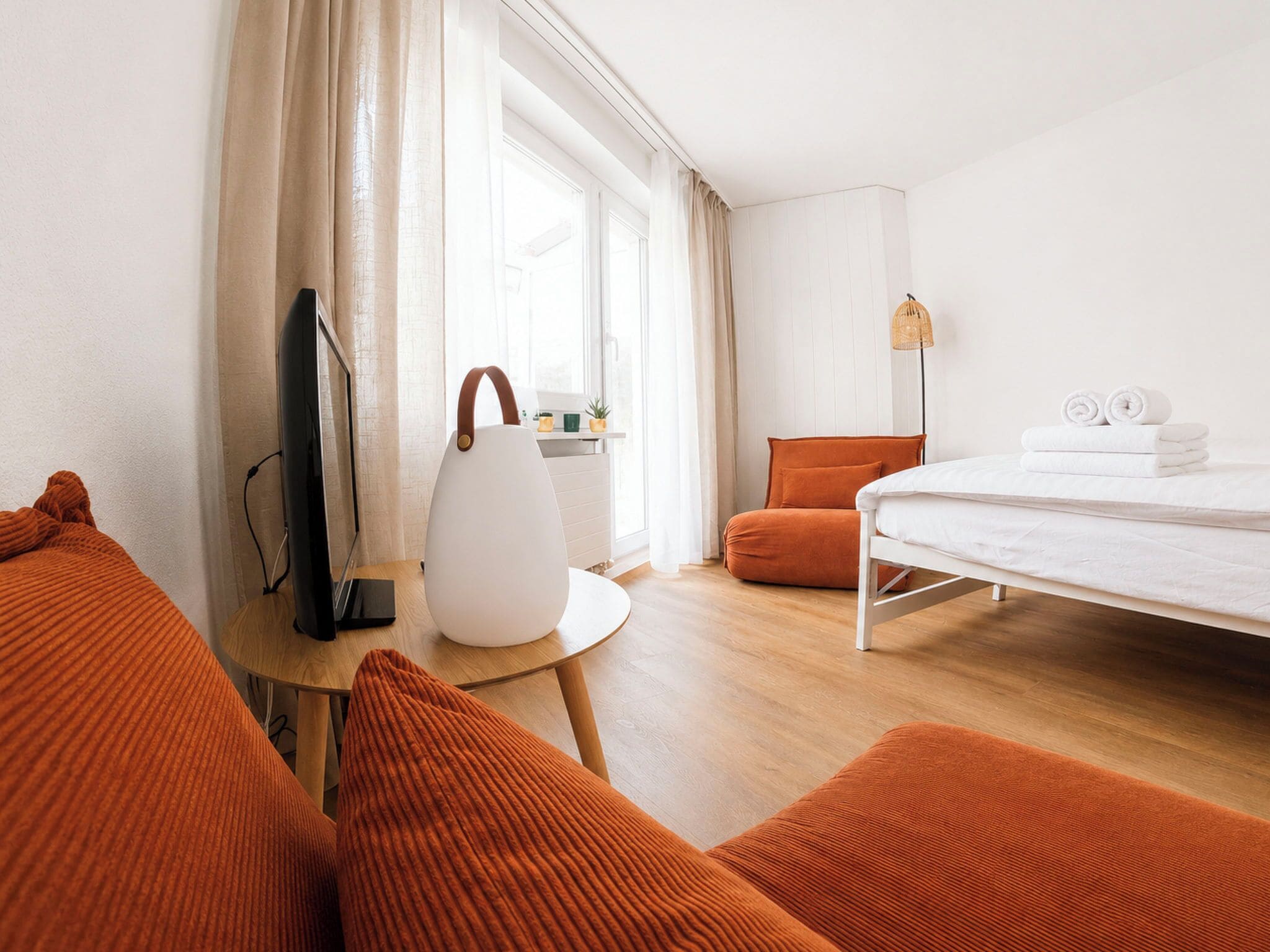Quarto de hotel com uma cama, uma escrivaninha com luminária, televisão e almofadas laranja.