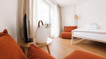Quarto de hotel com uma cama, uma escrivaninha com luminária, televisão e almofadas laranja.
