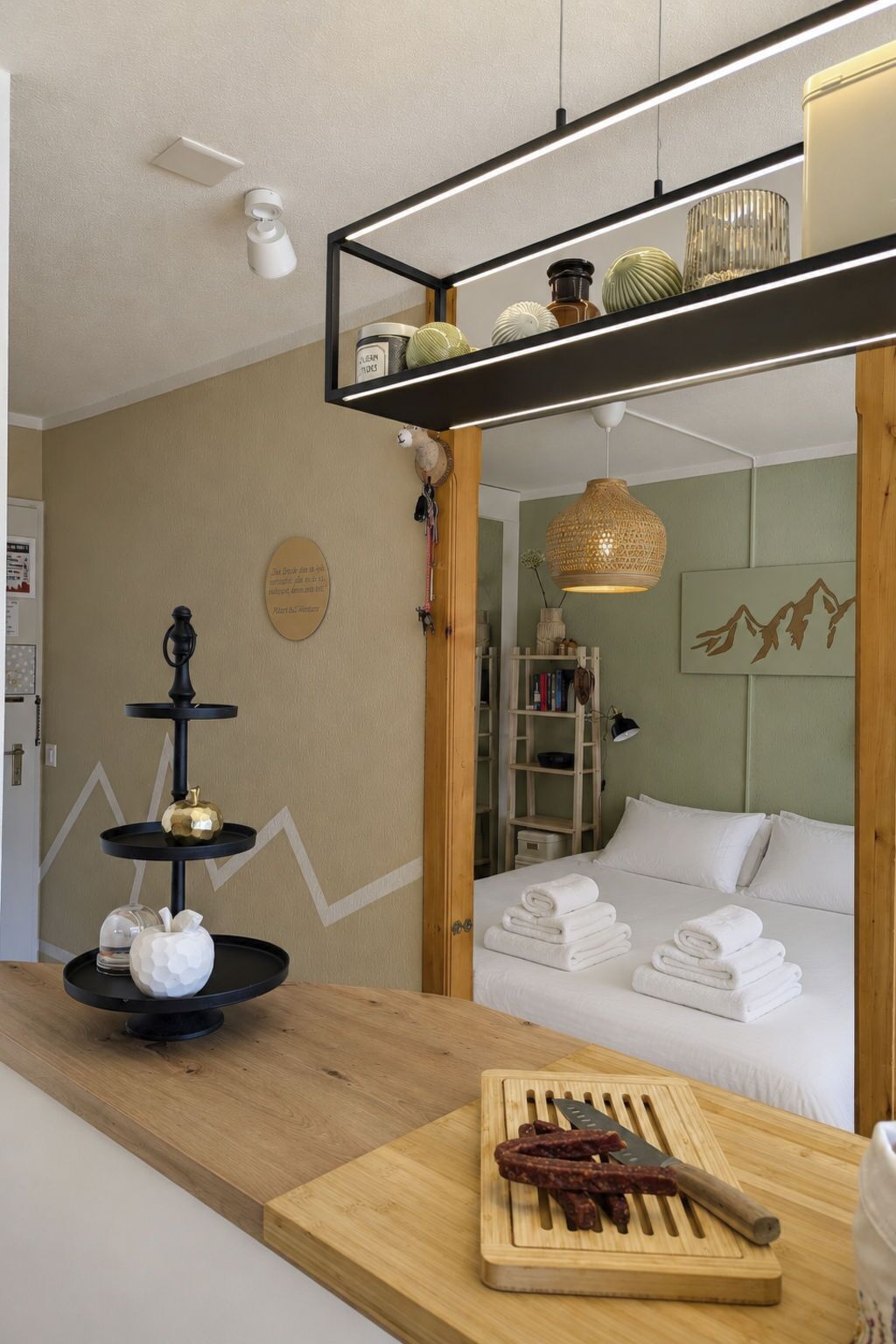 Une chambre d’hôtel avec un lit, une table en bois sur laquelle se trouvent une planche à découper et des saucisses, une étagère avec des objets décoratifs et une étagère murale avec des livres et une lampe.