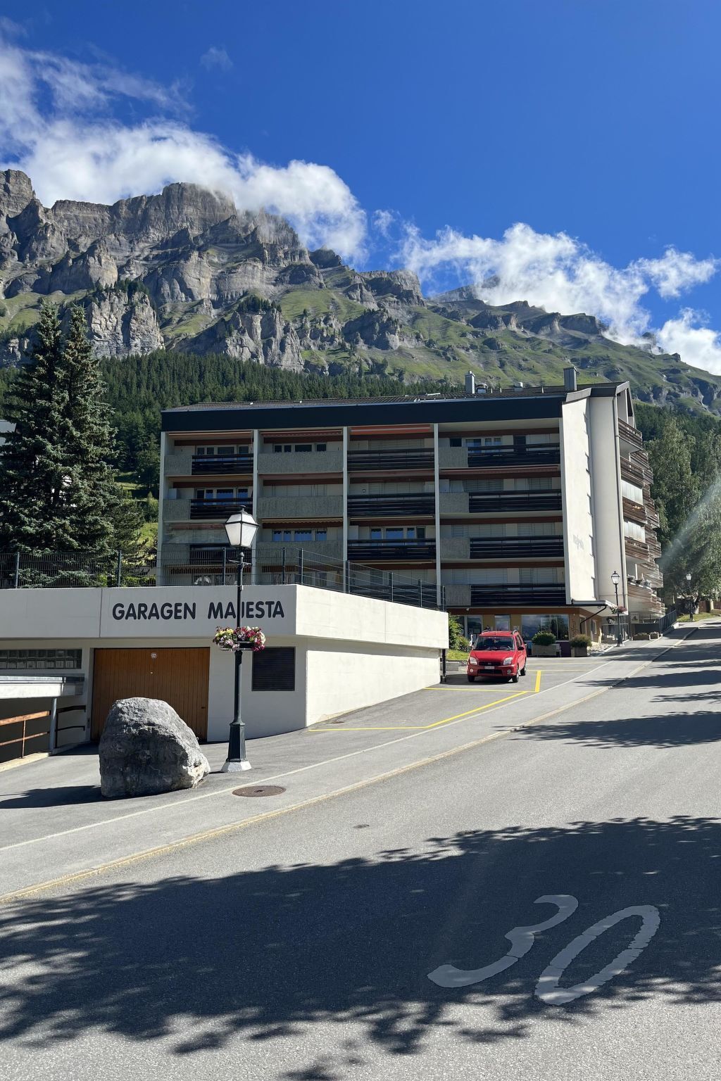 Um edifício com a placa 'GARAGEN MAISTETA', uma área de estacionamento e um carro vermelho estacionado em frente.