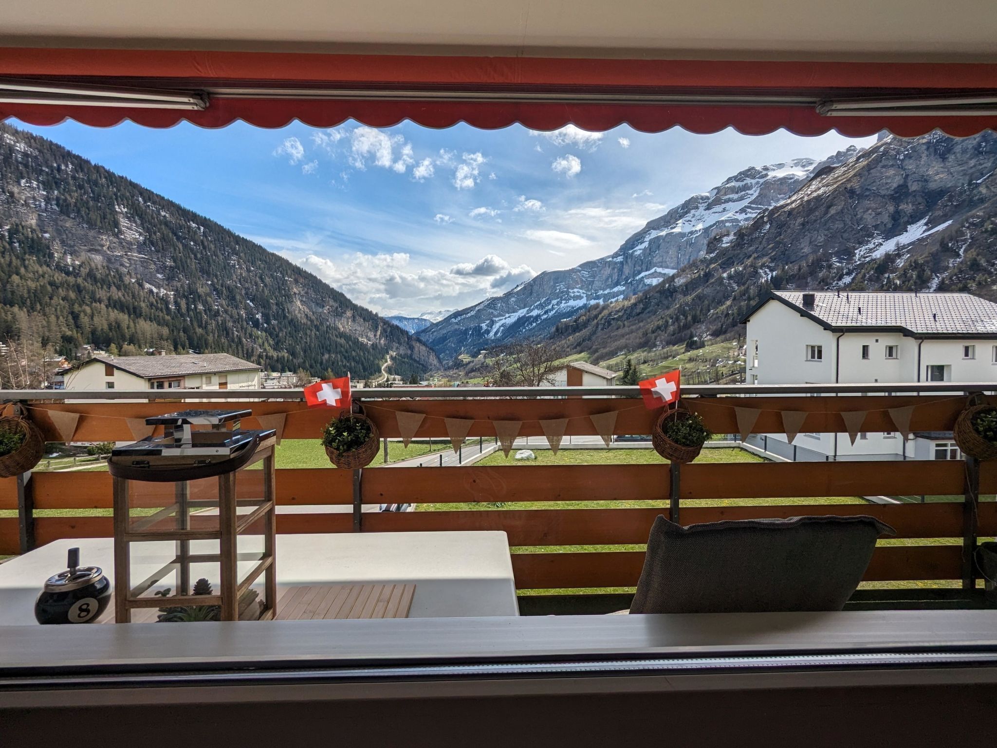 A balcony with a view of a valley and mountains, a table with a kettle, and two Swiss flags.