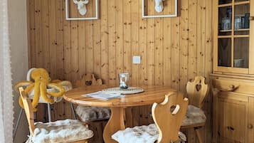 Um quarto com painéis de madeira, uma mesa redonda de madeira, quatro cadeiras com assentos acolchoados, uma luminária pendente branca e um armário com copos.