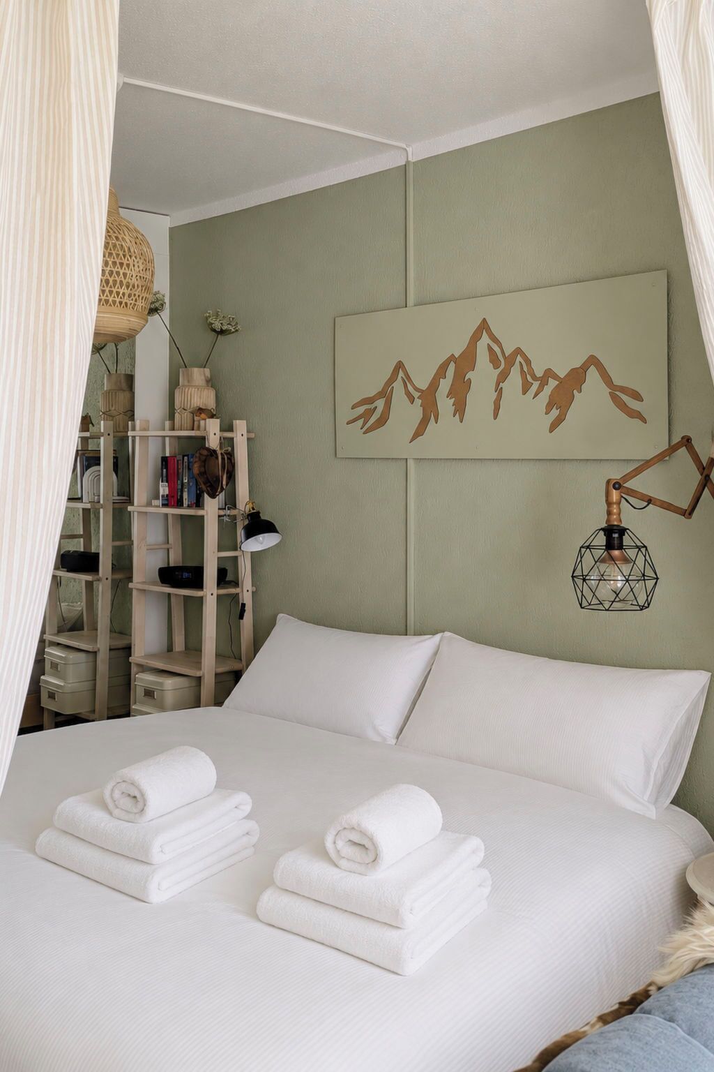 Un lit bien fait, avec du linge de lit blanc et des serviettes pliées, un luminaire géométrique fixé au mur et une œuvre d’art encadrée représentant une montagne.