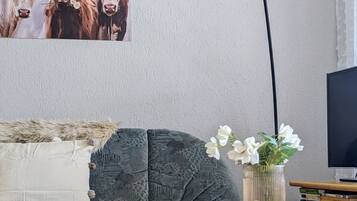 A living room with a grey sofa, a round white table with magazines, a vase with white flowers, and a painting of cows on the wall.