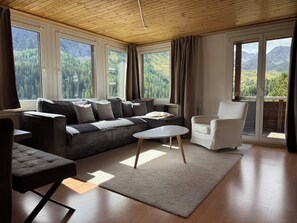 A modern living room with a sofa, a coffee table, and a chair. Large windows offer a view of a forested mountain range. - Miraflor Rosita by Arosa Holiday Appartement/Fewo, Dusche und Bad, WC, 2-Bettwoh by Interhome (Arosa)