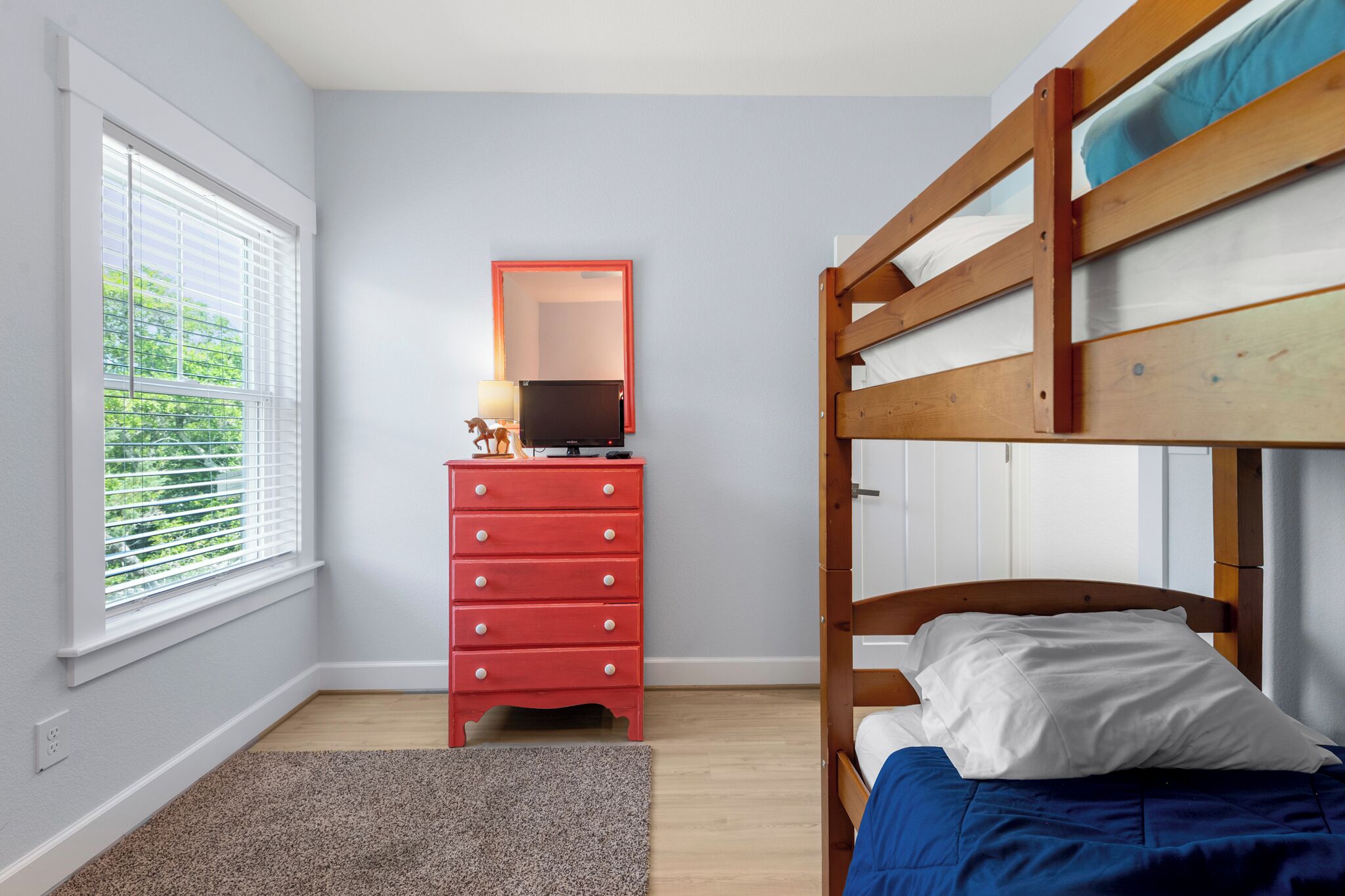 A bedroom with a bunk bed, a red dresser, a mirror, and a window with blinds.