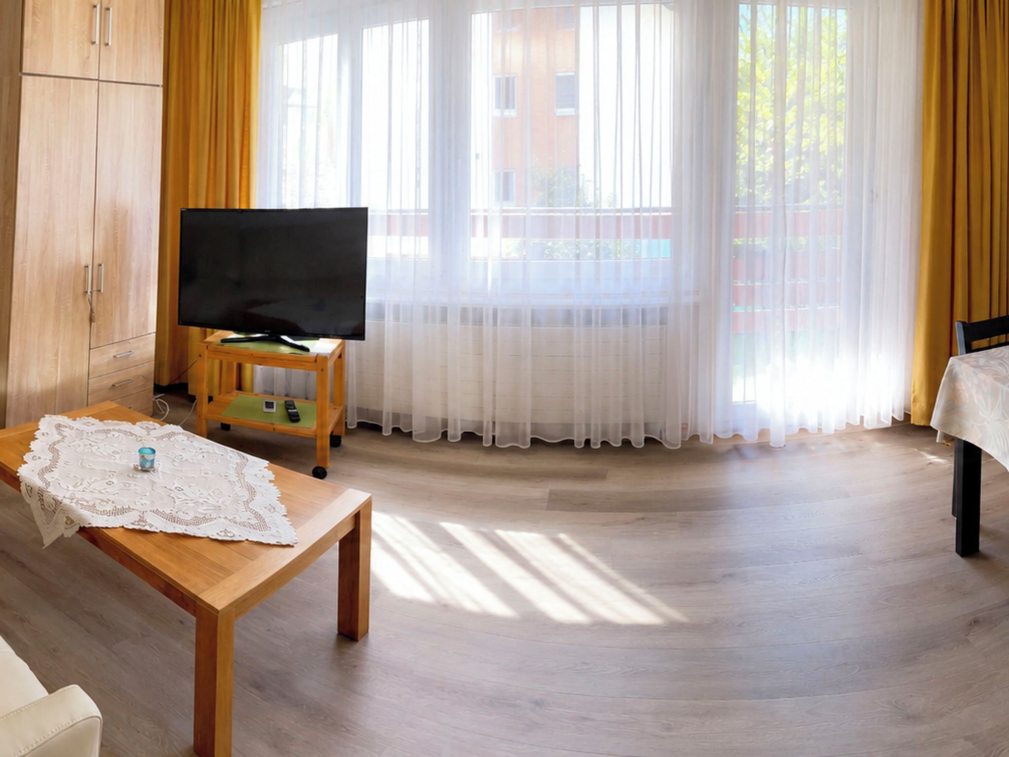 Una habitación con un armario de madera, un televisor sobre un mueble bajo, una mesa con mantel de encaje y una ventana con cortinas traslúcidas.