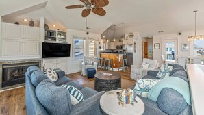 A spacious living room with a fireplace, a large sofa set, a round coffee table, and a kitchen area with bar stools. - NH3310: Chasing the Sun (Nags Head)