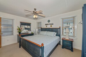 Une chambre à coucher comprenant un lit, une table de chevet, une commode, un ventilateur de plafond et une fenêtre avec des stores.