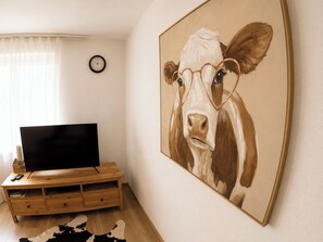 A living room with a flat-screen TV on a wooden TV stand, a cow painting on the wall, and a clock on the wall. - Majesta Leukerbad # 3-bed apartment, Dusche, WC by Interhome (Leukerbad)