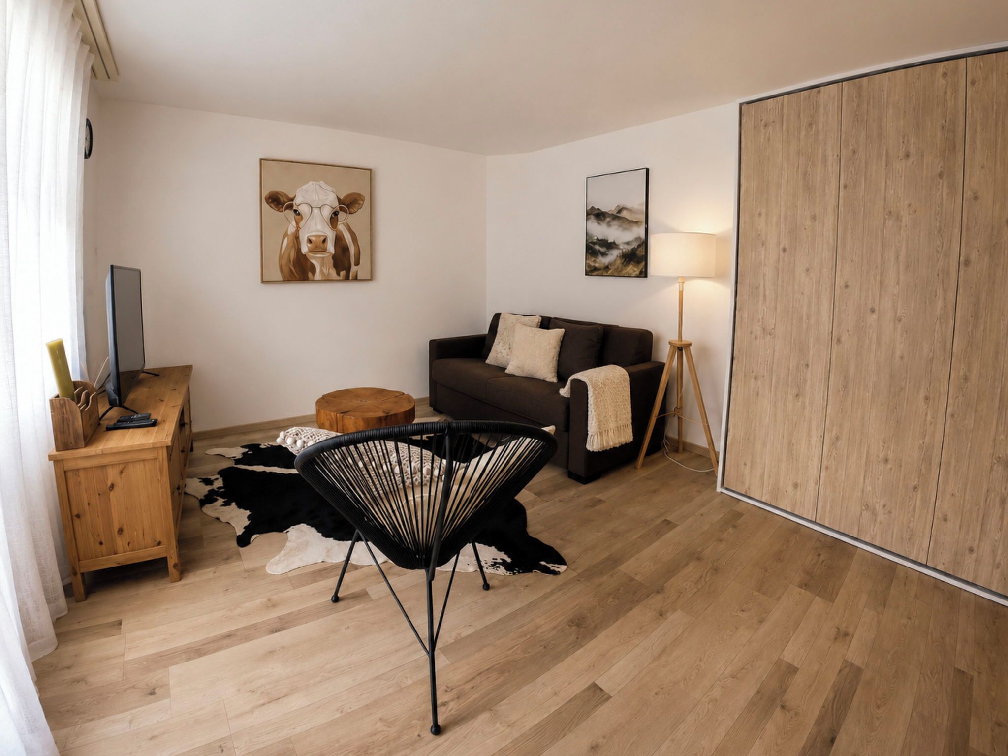 A modern living room with a wooden floor, a black chair, a brown sofa, a wooden desk, and a television.