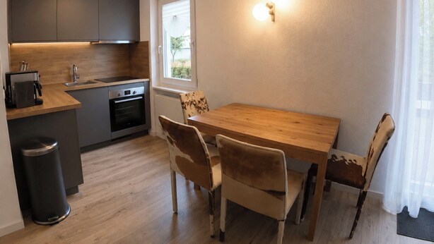 Una cucina compatta con zona pranzo, comprensiva di tavolo e sedie, cestino e finestra con tende.