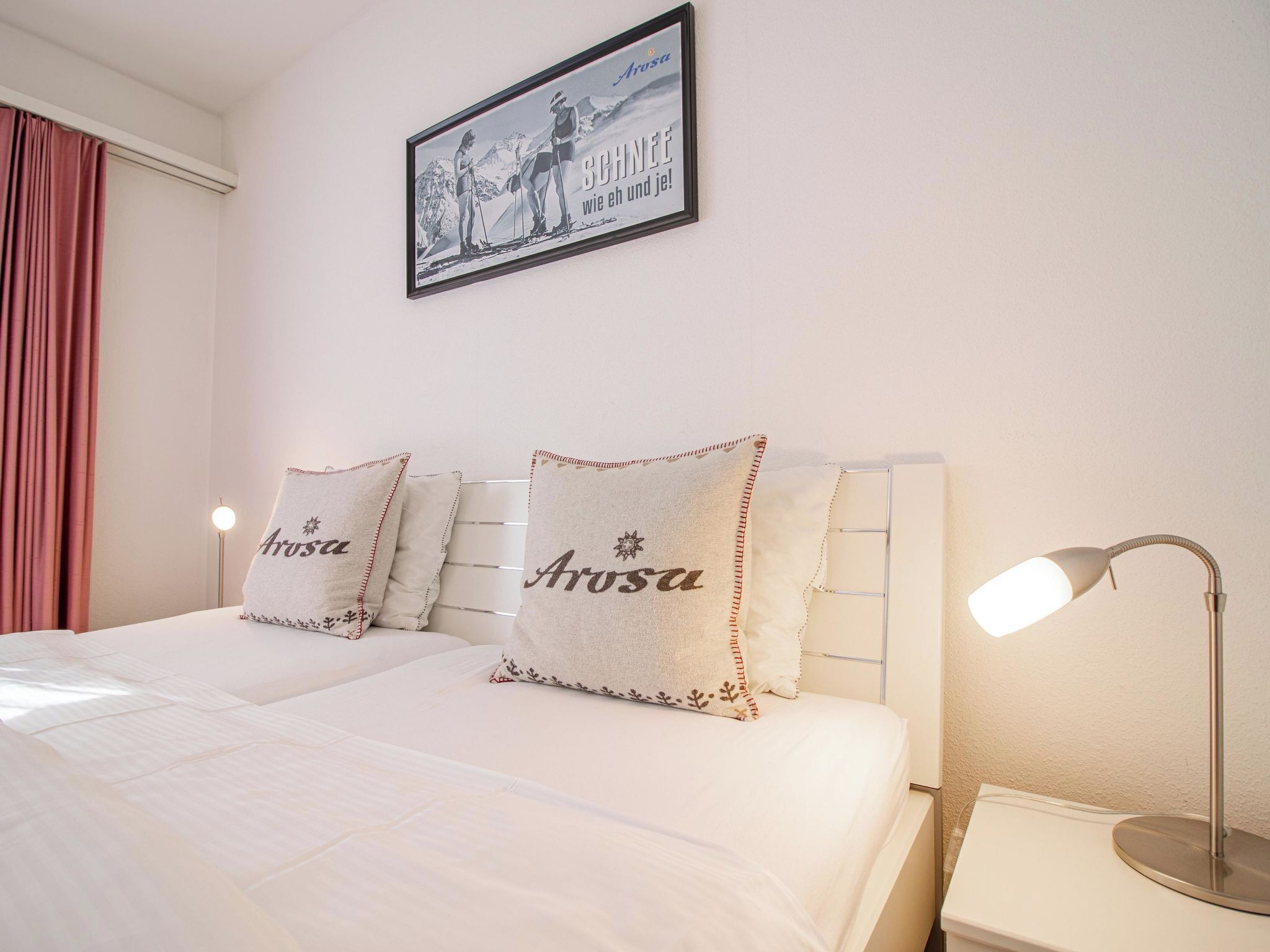 Een hotelkamer met een bed, twee kussens met de merknaam 'Arosa', een ingelijste foto aan de muur en een bedlampje.