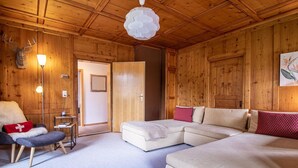 Et hyggeligt rum med en hjørnesofa, et lille bord og en stol. Loftet er træ, og der er en dør til et andet rum.