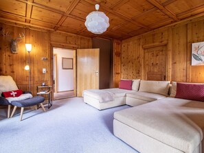 Ein gemütliches Zimmer mit einer Eckcouch, einem kleinen Tisch und einem Sessel. Holzdecke und eine Tür zu einem weiteren Raum.