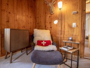 Ein gemütliches Zimmer mit Holzwänden, ein bequemer Sessel mit rotem Kissen, ein kleiner Tisch mit Kerze und eine Stehlampe.