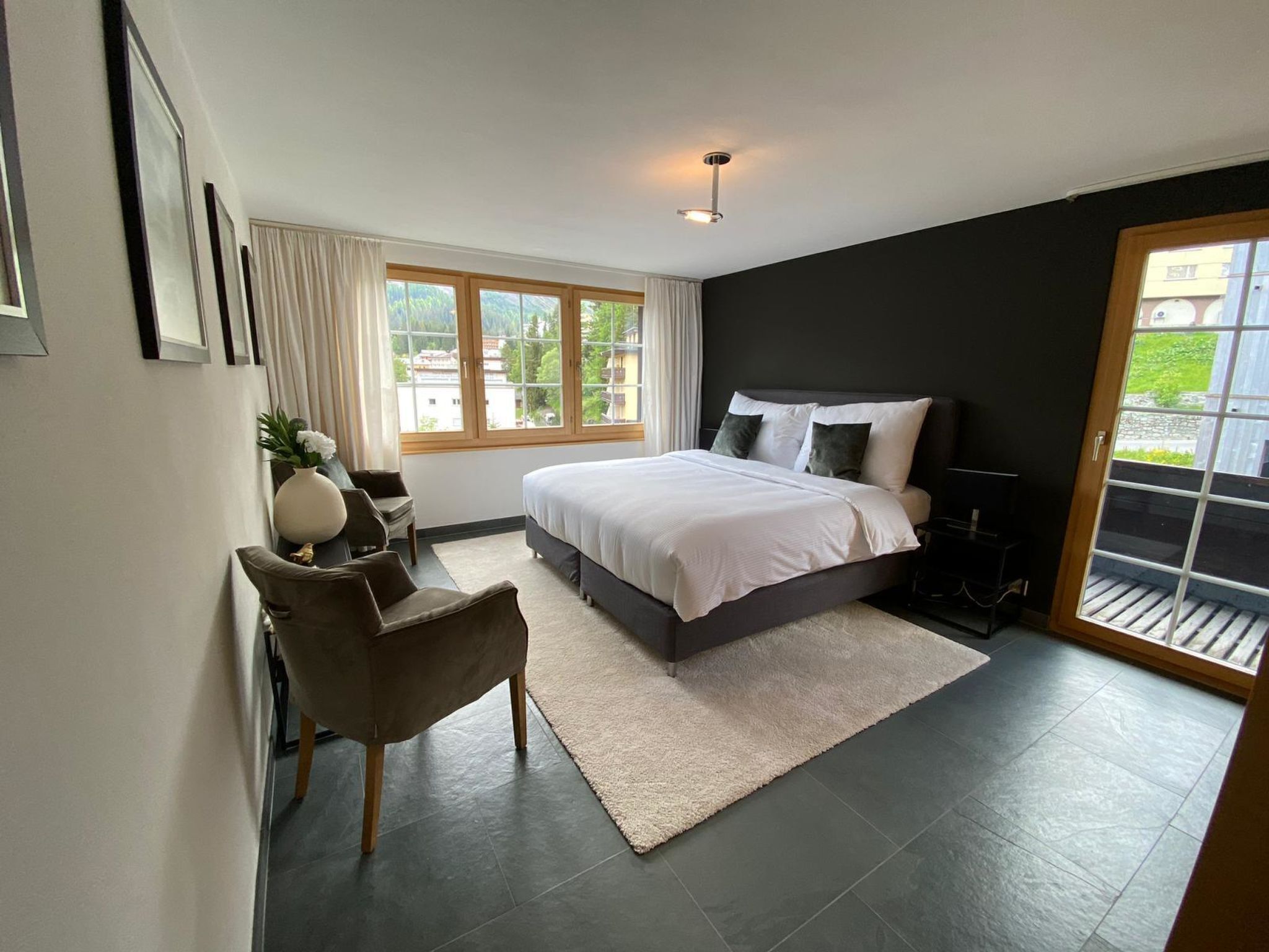 Quarto moderno com uma cama grande, uma escrivaninha e uma cadeira. Há uma janela com cortinas, uma porta que dá para uma área externa e uma luminária no teto.