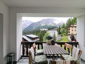 Un balcón con mesa y sillas, con vistas a un paisaje montañoso con edificios y árboles.